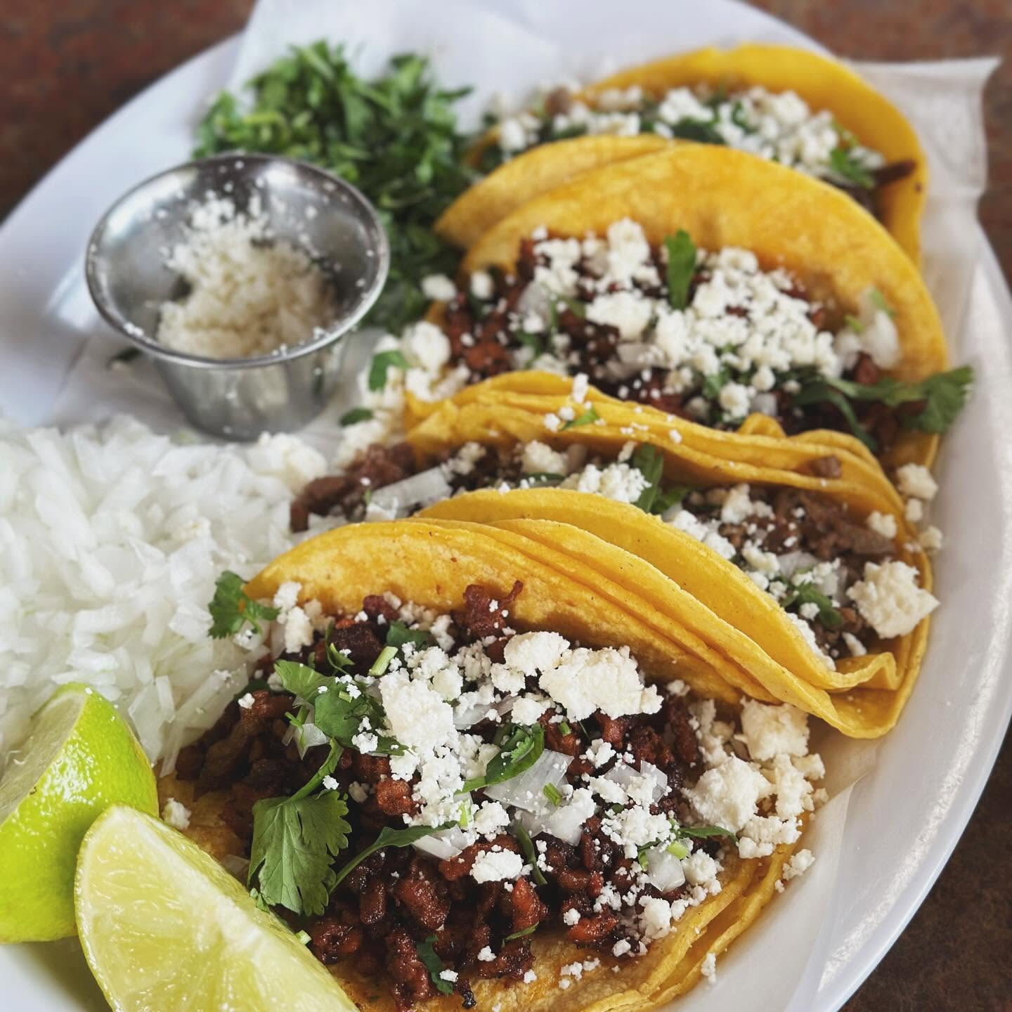 Three street tacos on yellow corn tortillas topped with cilantro, onion, and lime wedges.